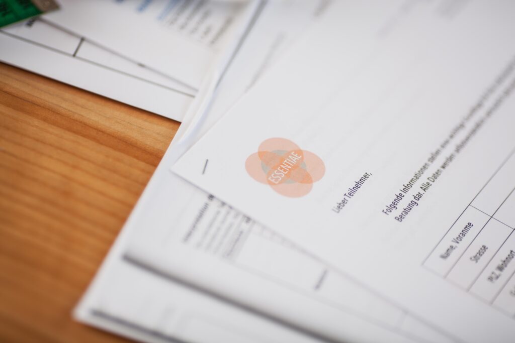 Ein Stapel ausgedruckter Dokumente liegt auf einem Holztisch. Das oberste Dokument trägt das Logo von "ESSENTIAE" in Orange und enthält eine Tabelle mit Namen und Zahlen. Der Fokus liegt auf dem oberen Teil des Papiers, während die restlichen Blätter leicht unscharf im Hintergrund liegen.